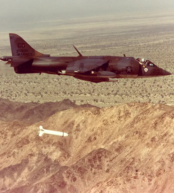 A Harrier AV-8A jet flies over a desert, releasing a missile toward the rocky terrain below.