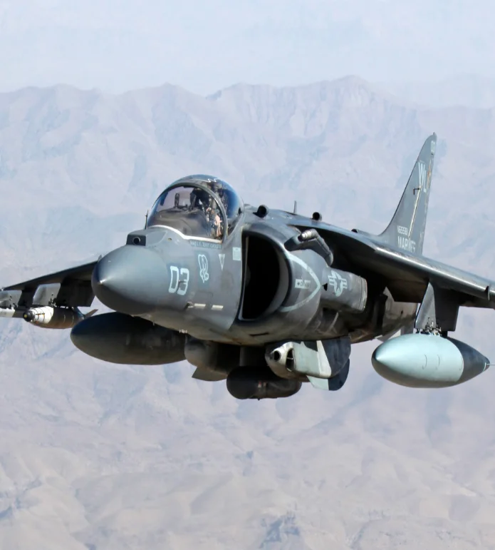 A Harrier AV-88 jet flies in the sky above mountains during daytime.