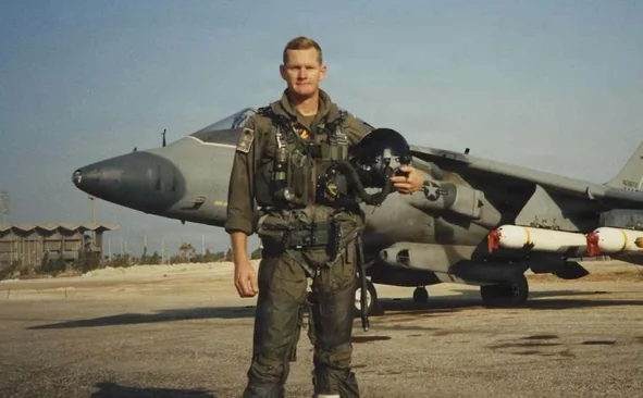Harrier Pilot in flight suit holding helmet stands in front of Harrier jet on an airfield under a clear sky.
