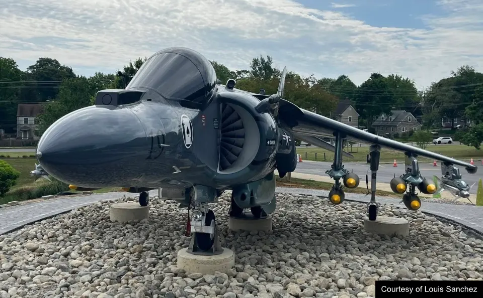A gray AV-8B Harrier II jet displayed outdoors on a rocky platform with trees and houses in the background.