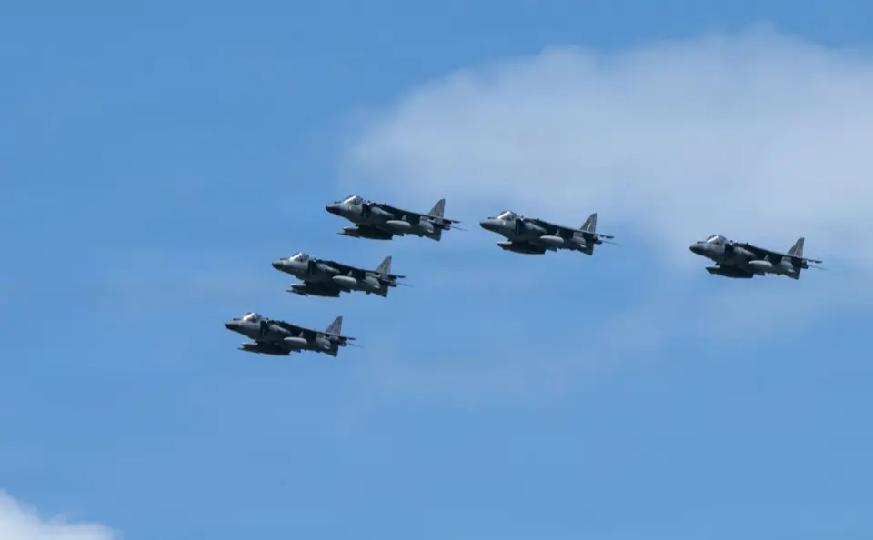 Five harrier jets are flying in formation against a blue sky with some clouds.
