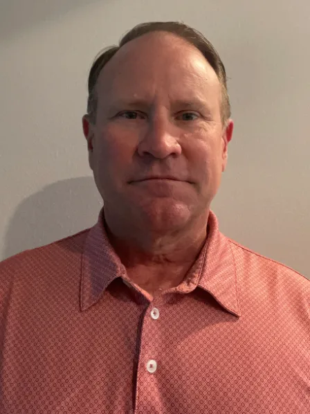Man in a red patterned collared shirt standing against a plain light-colored wall, facing the camera.
