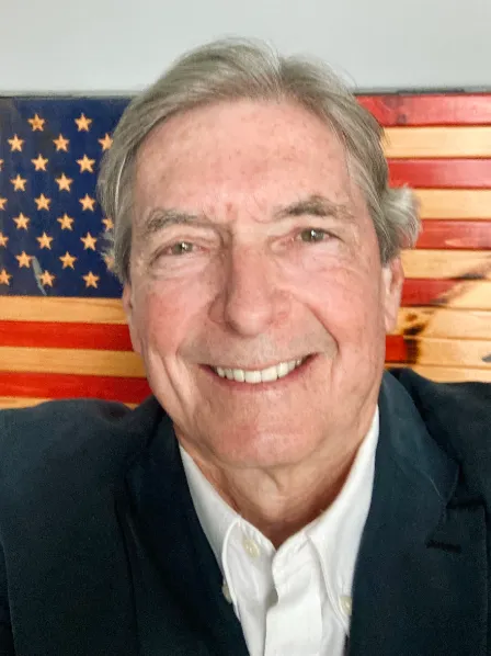 Smiling older man in a suit jacket, standing in front of a wooden American flag wall art.