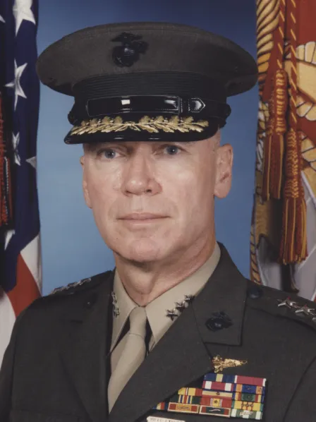 A U.S. military officer in uniform poses in front of American and service flags.