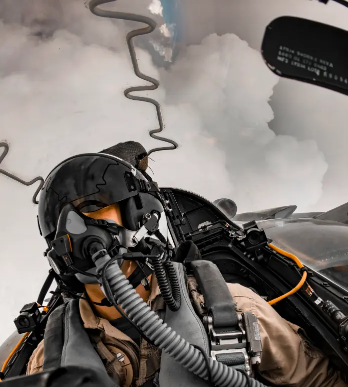 Pilot in a Harrier jet cockpit with oxygen mask; winding contrail path visible in the cloudy sky behind.
