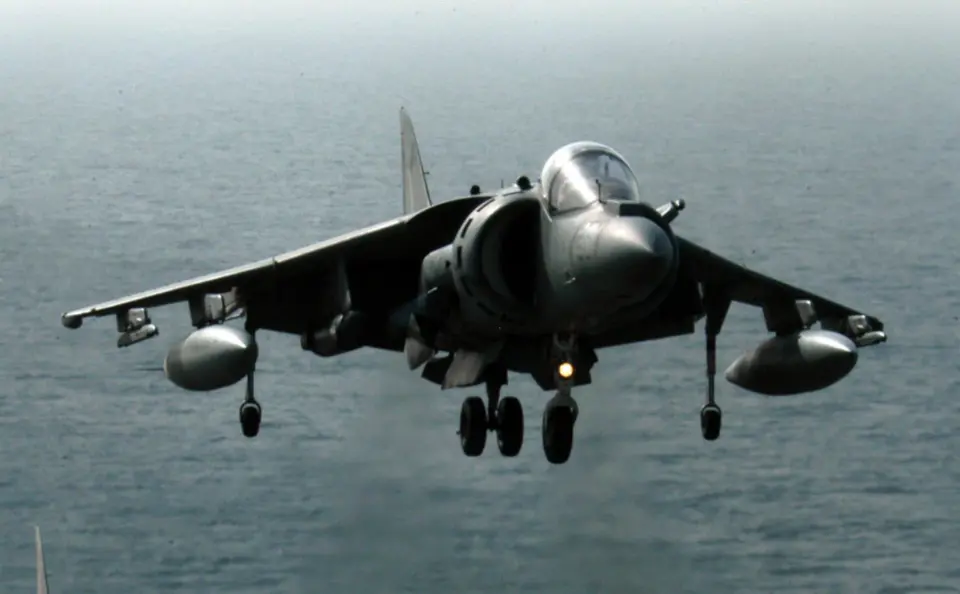 A Harrier jet hovers above the ocean, preparing to land with landing gear extended.