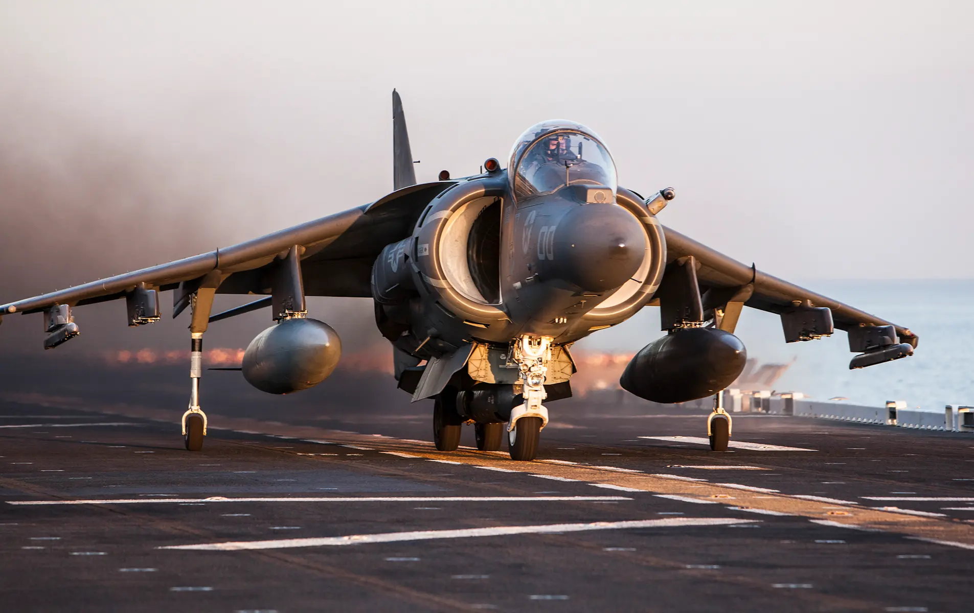 A Harrier jet takes off from an aircraft carrier with black smoke trailing behind it over the ocean.