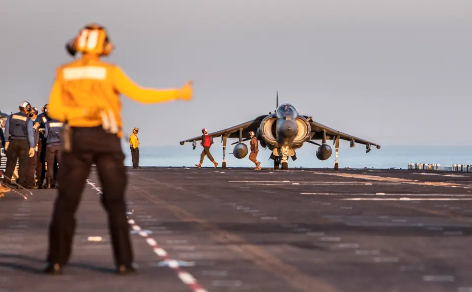 Person signals a Harrier jet landing on an aircraft carrier while crew members observe on the deck.