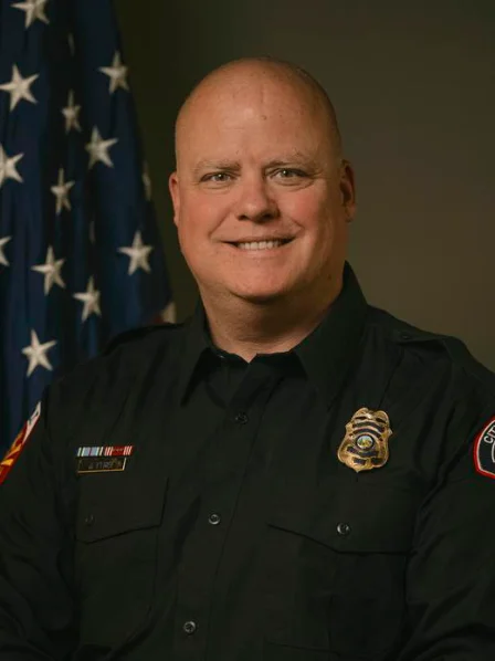 Jeremy tyree in a black uniform with badges, smiling, posed beside a U.S. flag.