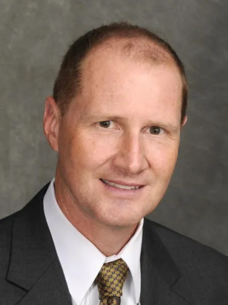 A man in a suit and tie smiles at the camera against a gray studio background.
