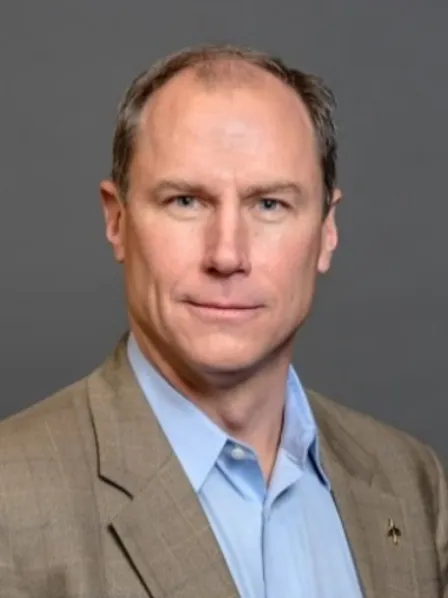A man in a tan suit jacket and blue shirt poses against a plain gray background, looking at the camera.
