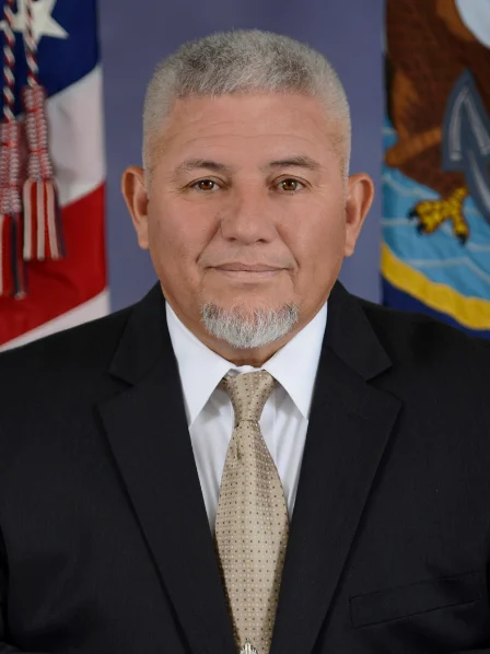 Pablo Sanchez in a suit and tie poses for formal portrait with flags in background.