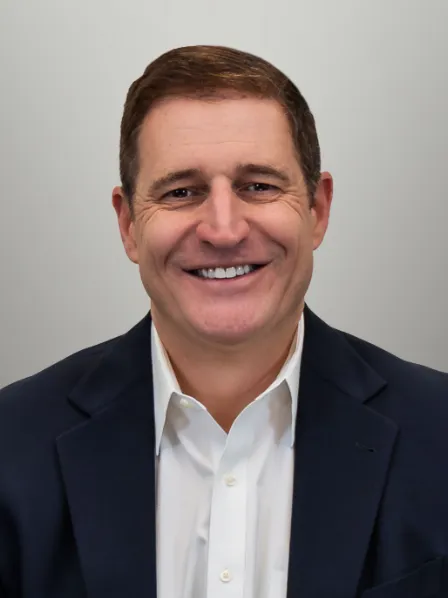 Smiling man in a dark blazer and white shirt poses in front of a plain light gray background.