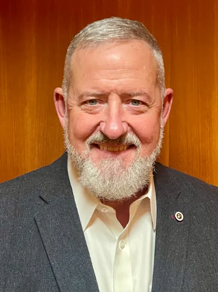Smiling man with short gray hair and a beard, wearing a dark suit jacket and white shirt, orange background.