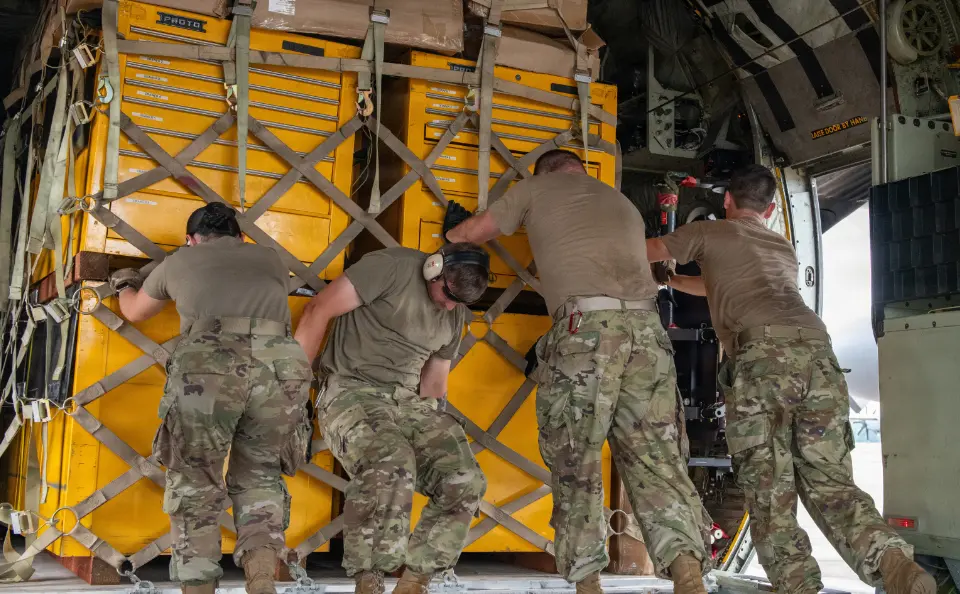 Four soldiers in camouflage uniforms push large yellow toolboxes inside a military cargo aircraft.