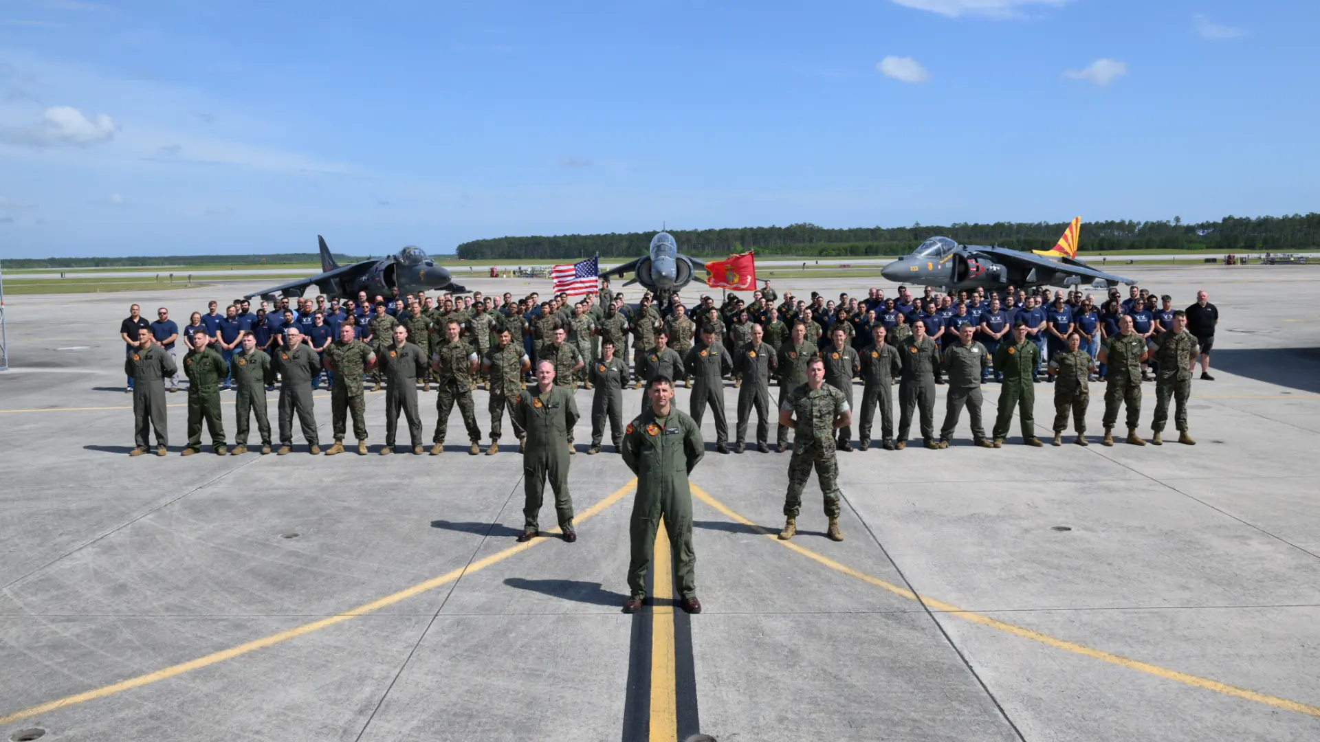 VMA-223 military personnel pose on runway in front of harrier jets, with flags visible behind them.