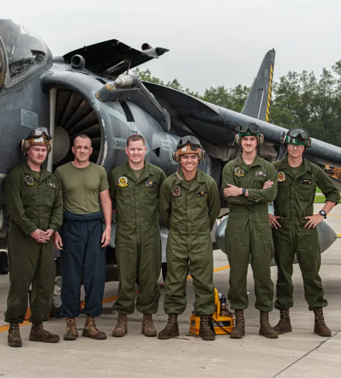 The Harrier Association Six pilots in green flight suits stand smiling in front of a military aircraft on a runway.