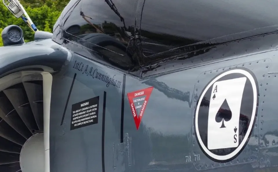 Close-up of a military aircraft with an Ace of Spades emblem and safety warning labels near the engine.